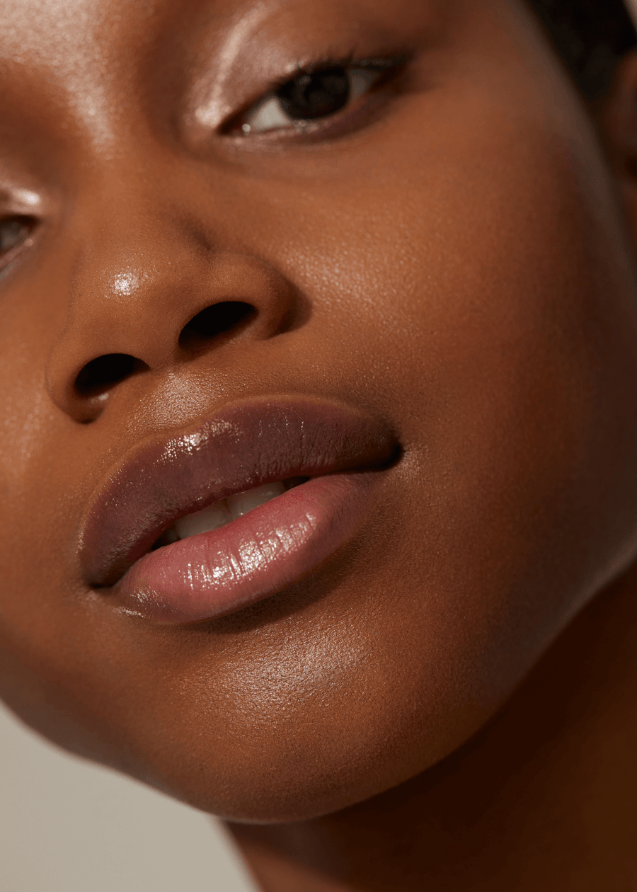 Woman with dark skin tone wearing a clear lip gloss in shade Dream from Minori